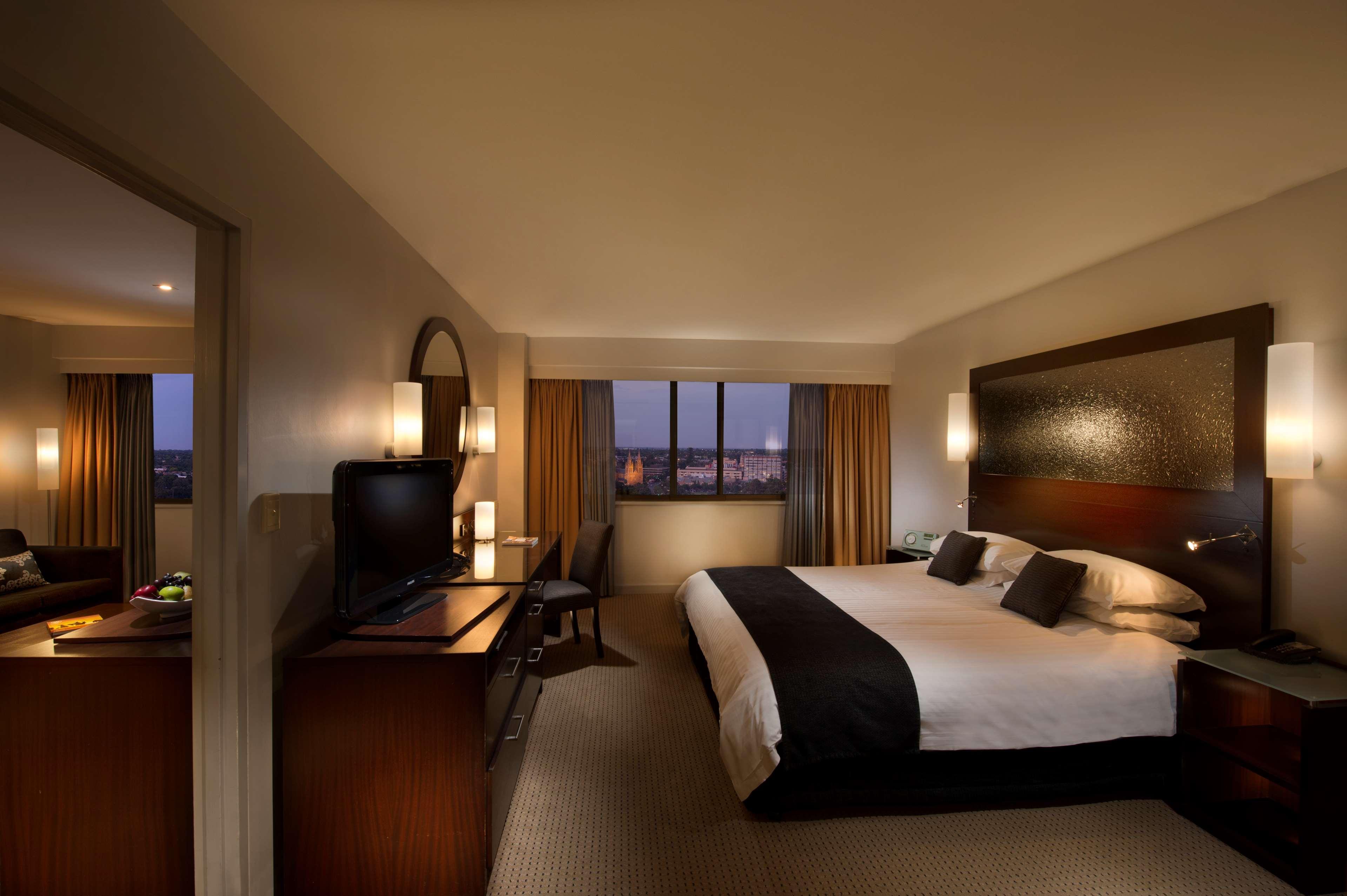 ホテル スタンフォード プラザ アデレード エクステリア 写真 The photo features a modern hotel room that includes a large bed with dark and light bedding, complemented by two pillows. The room is well-lit with warm lighting and has a contemporary aesthetic. To one side, there is a wooden dresser with a flat-sc