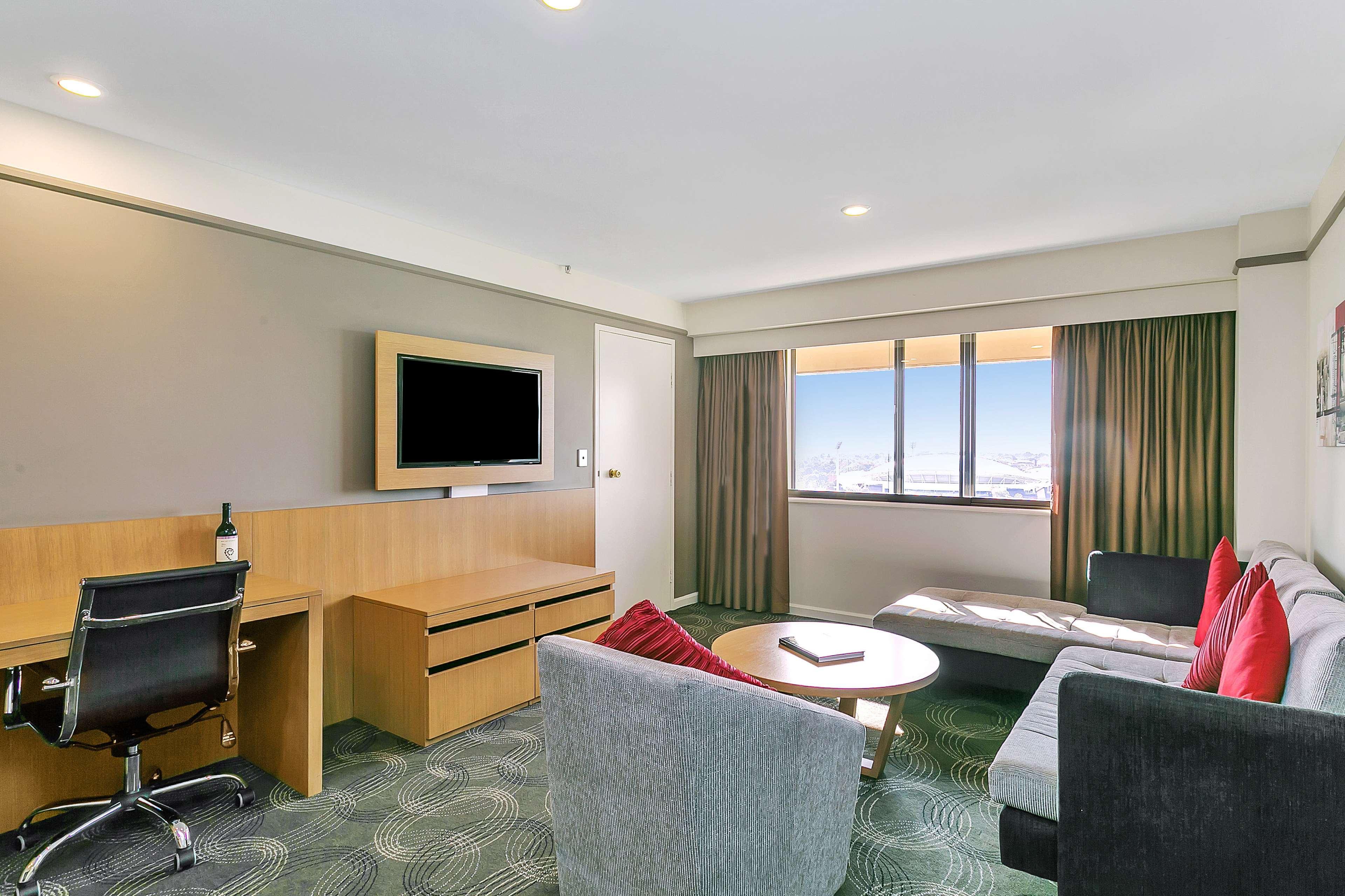 ホテル スタンフォード プラザ アデレード エクステリア 写真 The photo shows a modern hotel room or suite that features a cozy seating area. There is a comfortable sofa with red accent pillows, a round coffee table in the center, and a sleek desk with an office chair. On the wall, there is a flat-screen TV mou