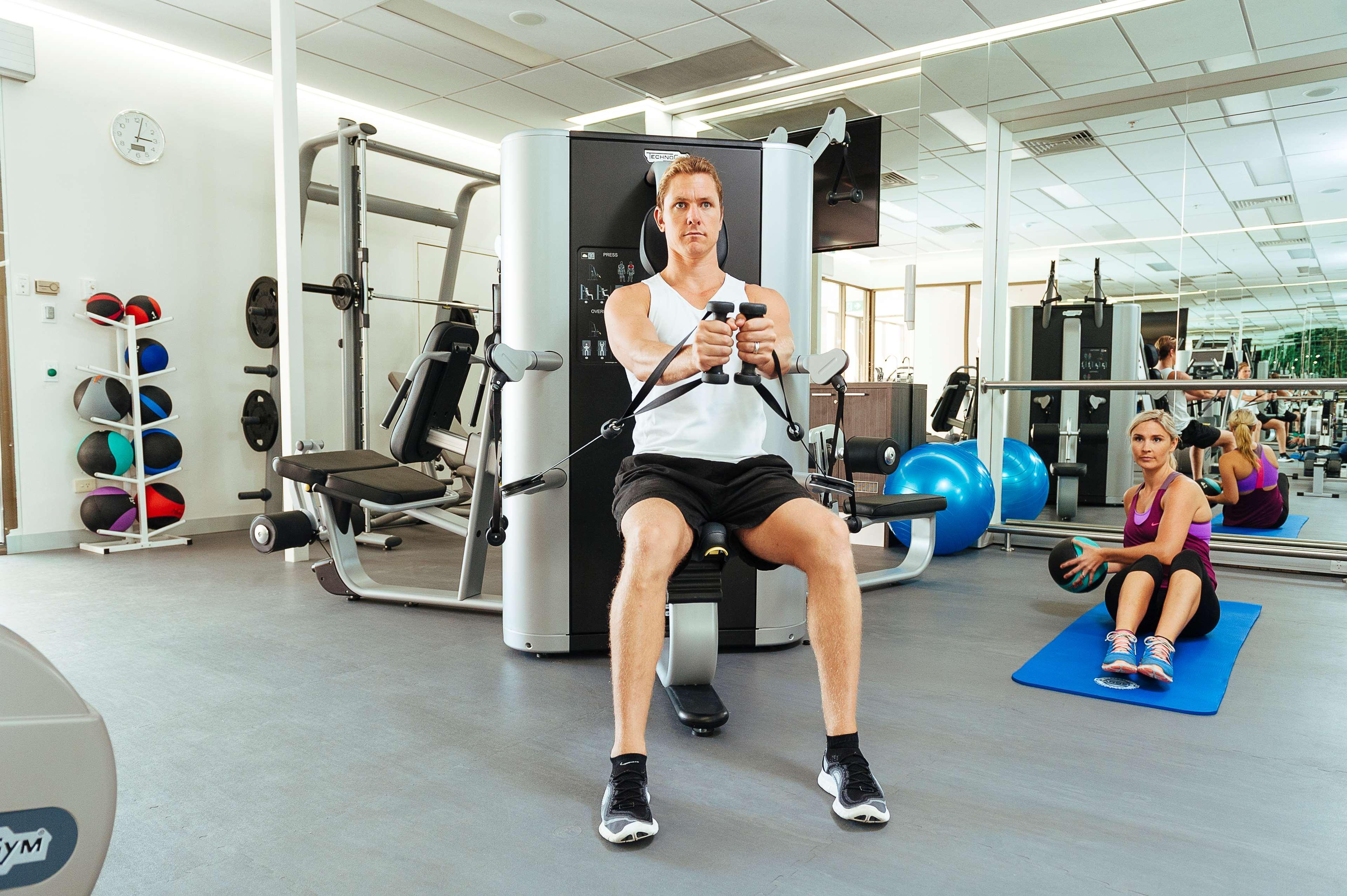 ホテル スタンフォード プラザ アデレード エクステリア 写真 The photo shows a gym setting with two individuals engaged in workout activities. 

In the foreground, a man is seated on a machine, using the handles to perform a rowing exercise. He is wearing a sleeveless shirt and shorts, indicating an active wor