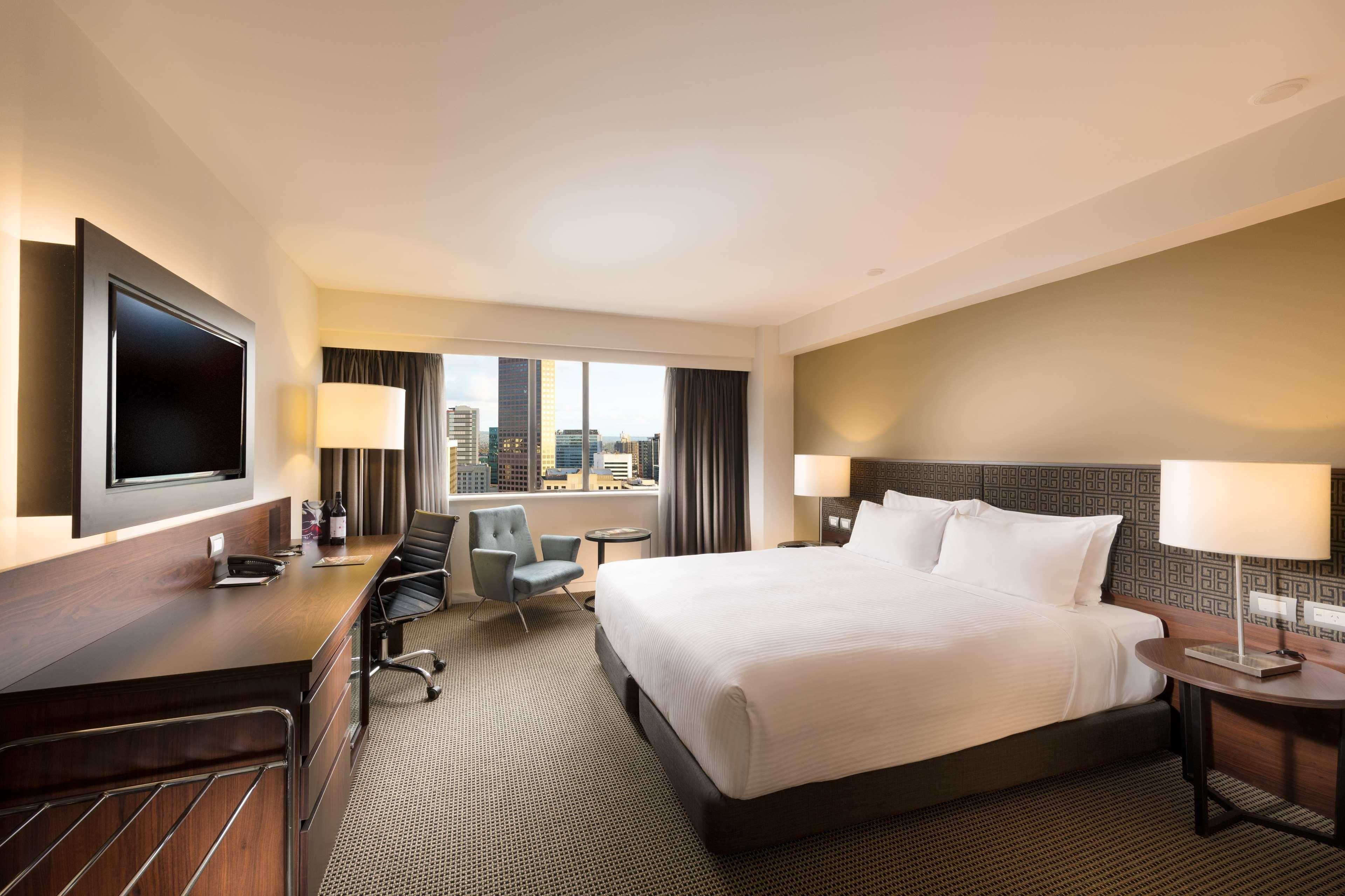 ホテル スタンフォード プラザ アデレード エクステリア 写真 The photo shows a modern hotel room with a contemporary design. It features a large bed with white linens, a bedside table on either side with lamps, and a dark wood accent wall. There's a work desk with an office chair and a flat-screen television m