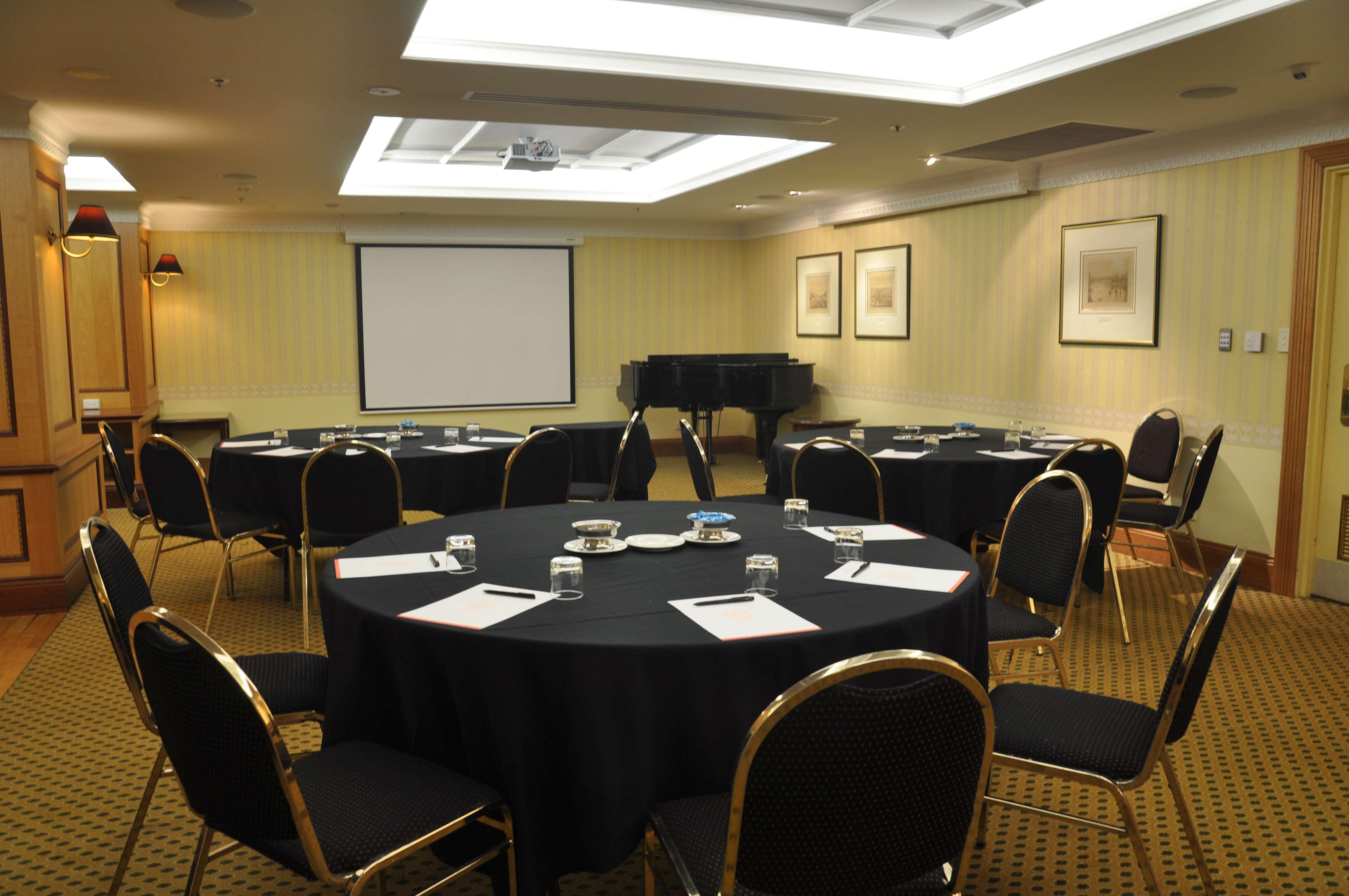 ホテル スタンフォード プラザ アデレード エクステリア 写真 The photo shows a conference room set up for a meeting or presentation. There are several round tables covered with black tablecloths, each surrounded by black upholstered chairs. On the tables, there are notepads and glasses of water, indicating tha