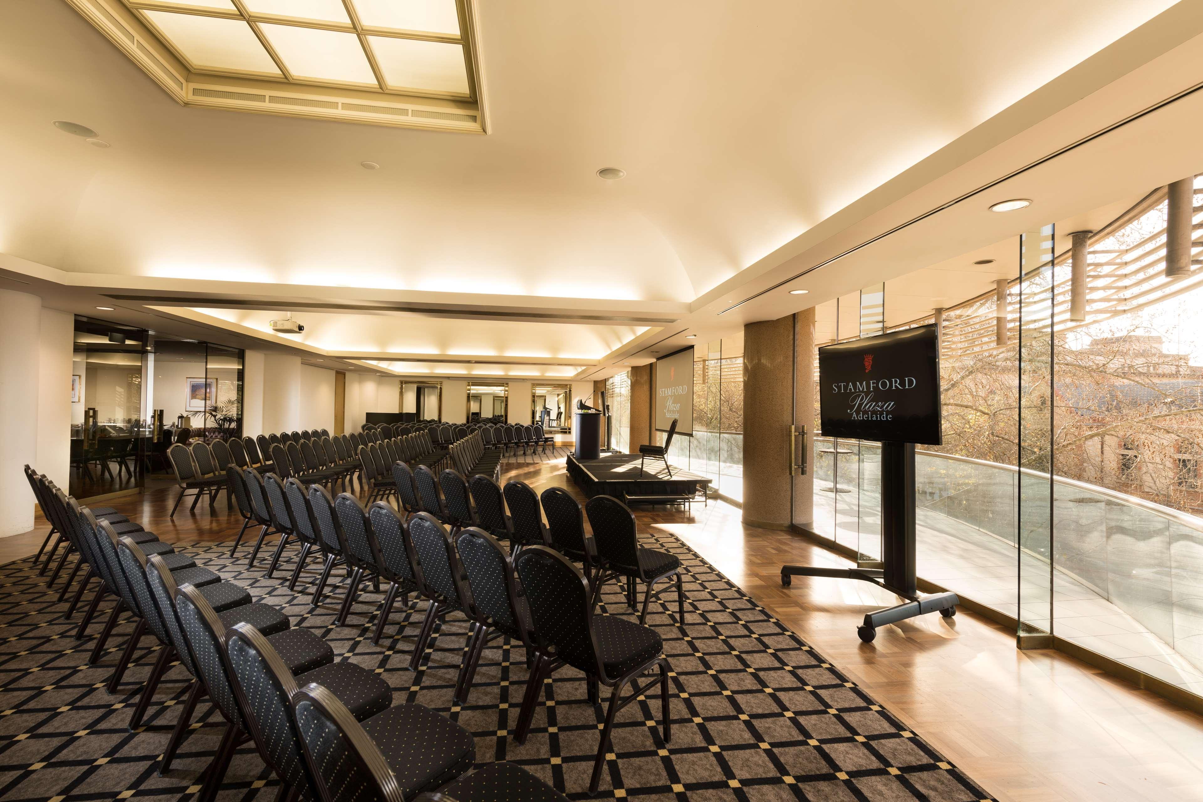 ホテル スタンフォード プラザ アデレード エクステリア 写真 The photo shows a large, elegantly designed event space or meeting room. There are rows of black chairs set up facing a presentation area. The flooring features a patterned carpet, and large windows allow natural light to enter the room. On one side,