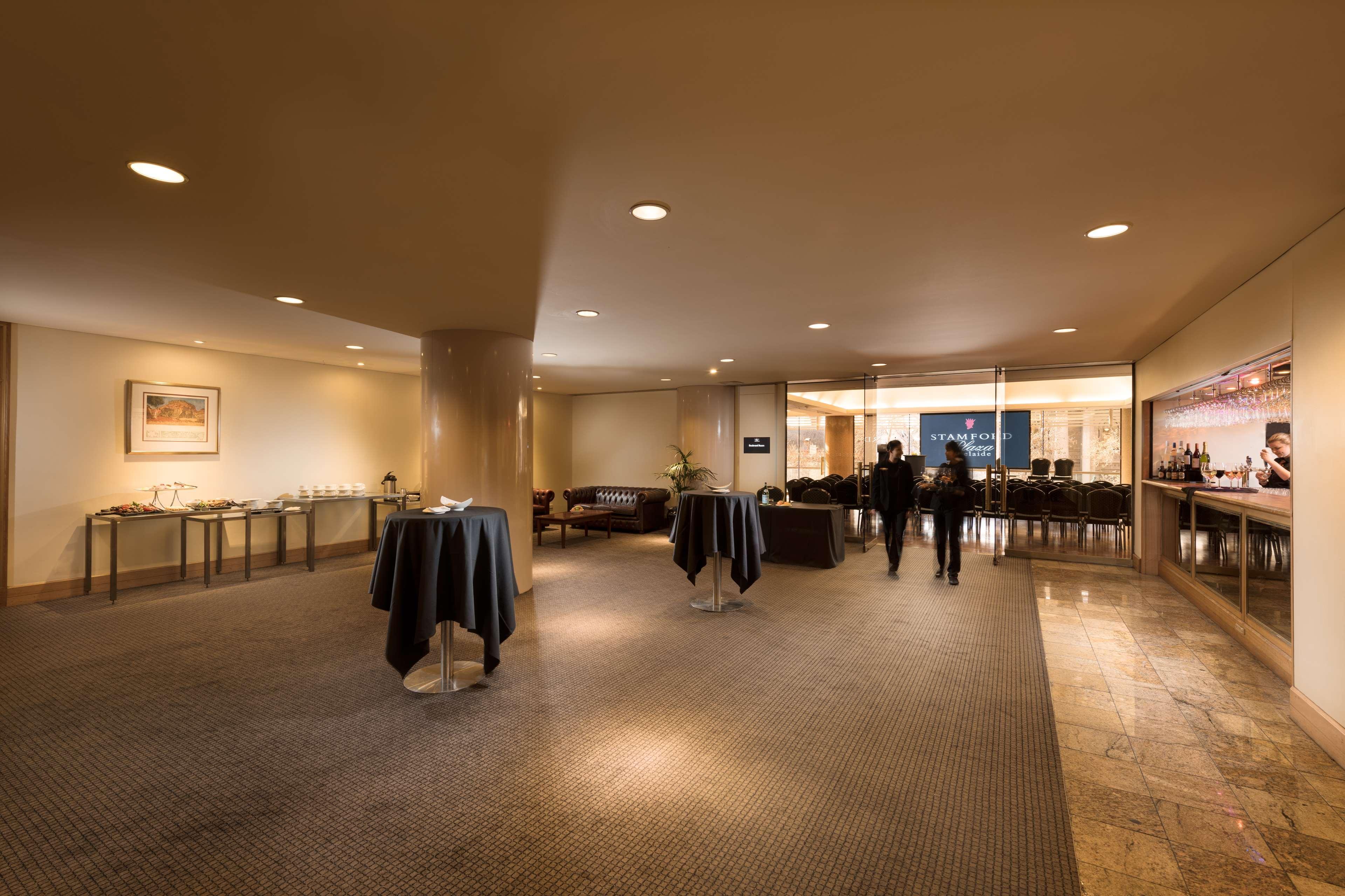 ホテル スタンフォード プラザ アデレード エクステリア 写真 The image shows an indoor space that appears to be a lounge or reception area. There are several round tables with dark tablecloths positioned throughout the room. The floor is carpeted, and the lighting is warm and somewhat dim. In the background, t