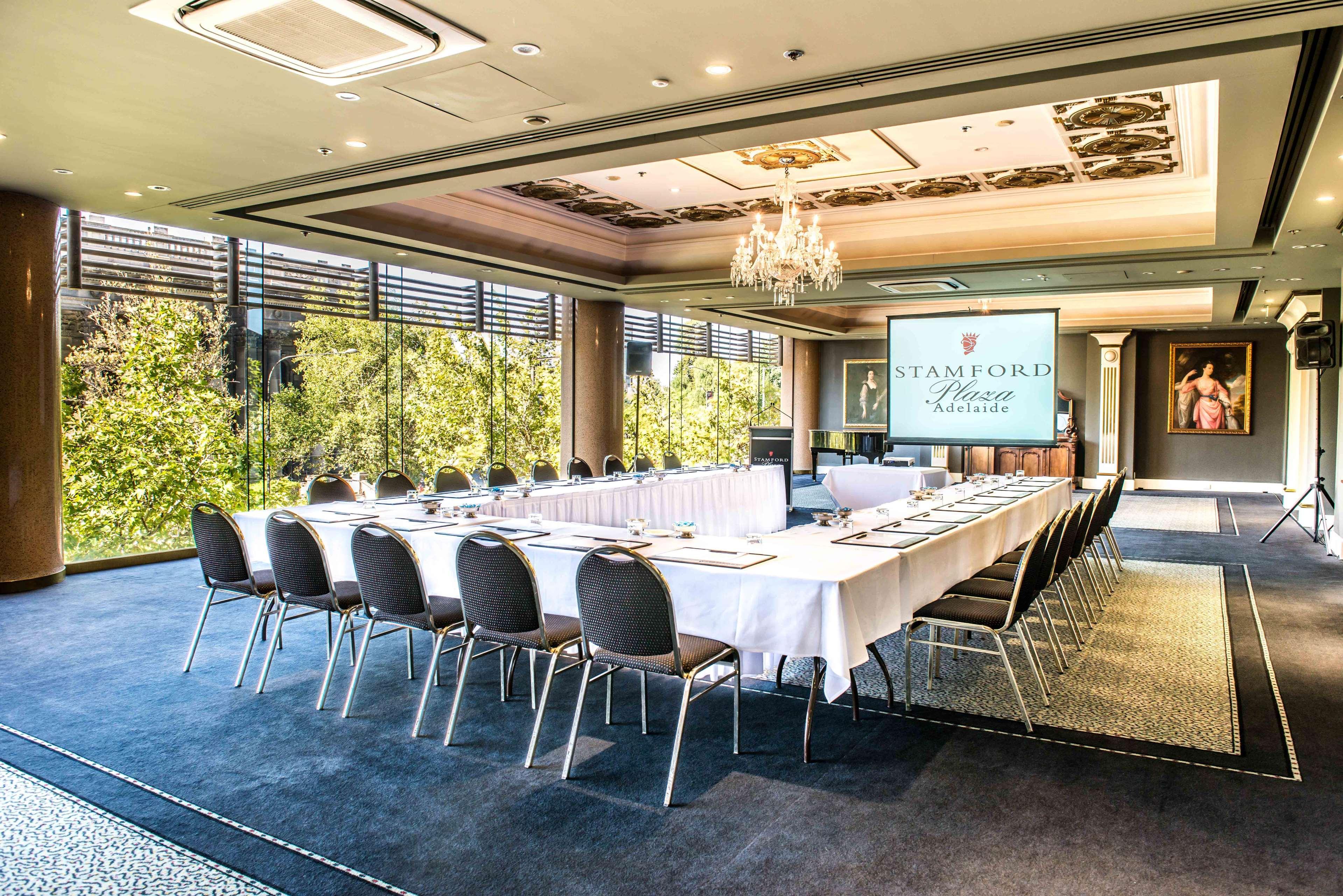 ホテル スタンフォード プラザ アデレード エクステリア 写真 The photo shows a stylish conference room set up for a meeting or event. There is a long rectangular table covered with a white tablecloth, surrounded by black chairs. A large screen displays the name "Stamford" along with a logo. The room features l