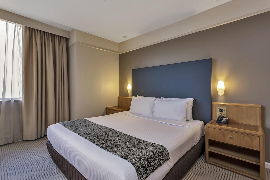 ホテル スタンフォード プラザ アデレード エクステリア 写真 The photo shows a modern hotel room. It features a double bed with a white duvet and a decorative grey and black throw. The headboard is a solid dark color. There are two bedside tables on either side of the bed, each with a lamp. Natural light enter